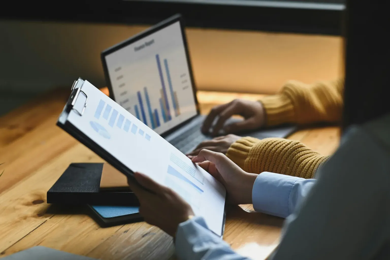 Two people reviewing printed charts beside a laptop displaying a bar chart